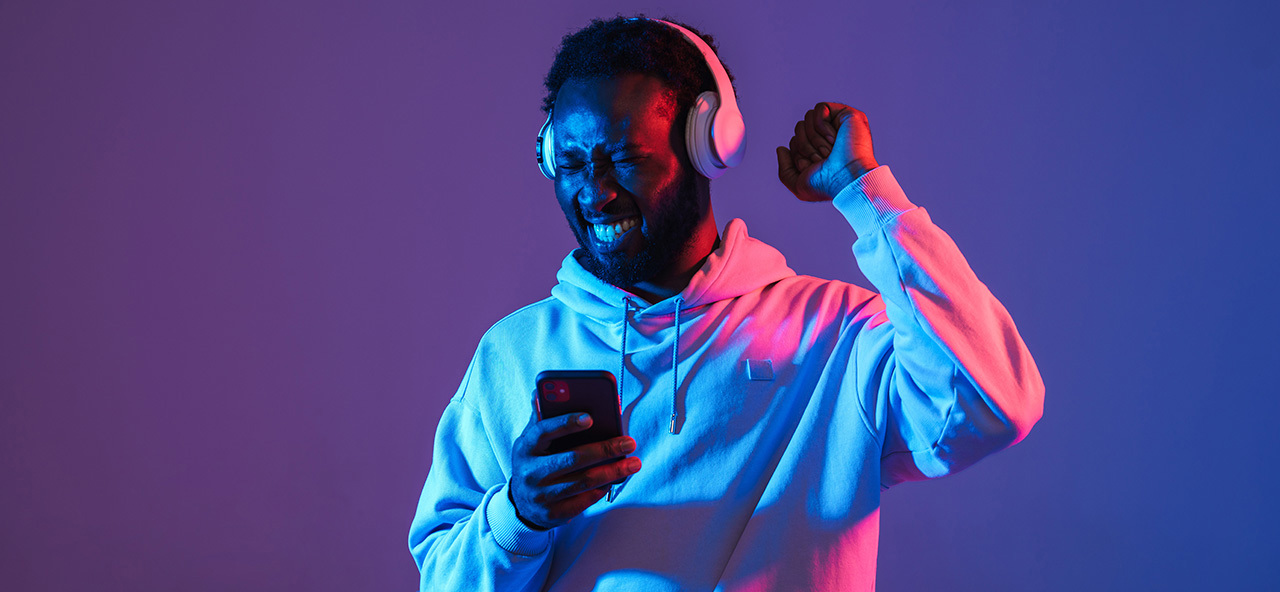 Man med hörlurar som ler och firar medan han tittar på sin mobil, upplyst av blått och rosa ljus.