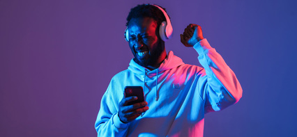 Man med hörlurar som ler och firar medan han tittar på sin mobil, upplyst av blått och rosa ljus.