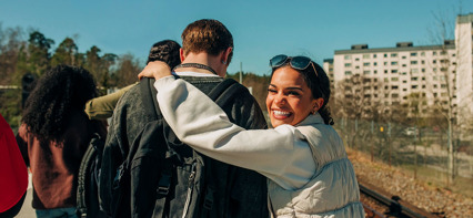 Leende kvinna kramar en vän i en grupp som går utomhus i solen.